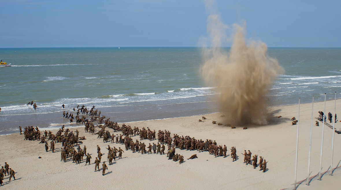 Actors dressed as WWII soldiers fight on a Dunkirk beach. A big explosion creates a cloud of sand on the beach